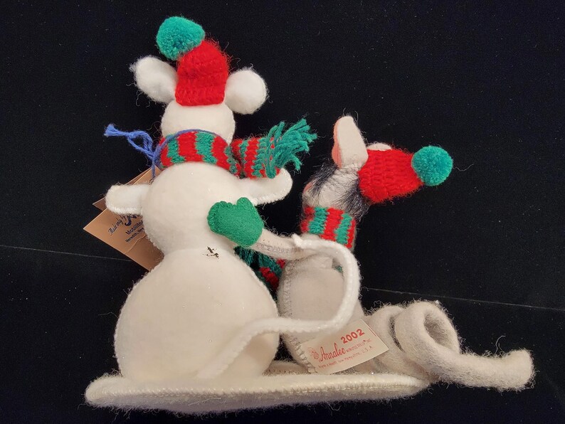 Puede incluir: Una caprichosa figura de mu&ntilde;eco de nieve con una bufanda a rayas rojas y verdes, un gorro a juego y guantes verdes. El mu&ntilde;eco de nieve est&aacute; hecho de fieltro blanco y tiene una larga cola rizada. Una etiqueta dice "2002" y "Amalie".