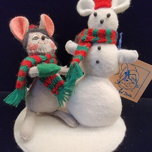 Puede incluir: Una decoraci&oacute;n navide&ntilde;a caprichosa con dos ratones de fieltro. Un rat&oacute;n lleva una bufanda roja y verde y un guante verde. El otro rat&oacute;n est&aacute; encima de un mu&ntilde;eco de nieve con una bufanda roja y verde y un gorro rojo. La base es un c&iacute;rculo de fieltro blanco.