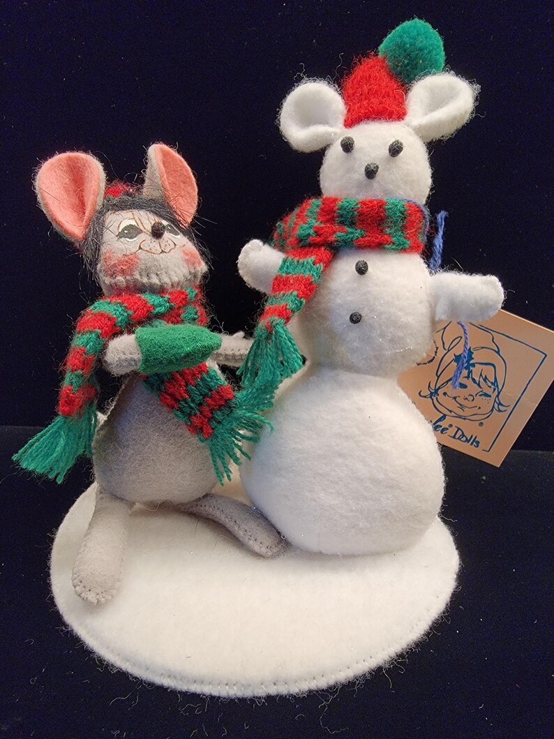 Puede incluir: Una decoraci&oacute;n navide&ntilde;a caprichosa con dos ratones de fieltro. Un rat&oacute;n lleva una bufanda roja y verde y un guante verde. El otro rat&oacute;n est&aacute; encima de un mu&ntilde;eco de nieve con una bufanda roja y verde y un gorro rojo. La base es un c&iacute;rculo de fieltro blanco.