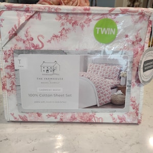 May include: A Twin-size 100% cotton sheet set in a clear plastic package. The sheets have a white background with a pink floral pattern. The packaging includes the text "THE FARMHOUSE" and "GARMENT WASH".