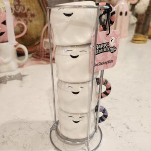 May include: Set of four white ghost-shaped ceramic mugs with black eyes and smiling mouths. The mugs are stacked on a silver metal stand. The mugs have pink and black striped handles. A pink and white label reads "Happy Hauntings 8oz Stacking Cups".