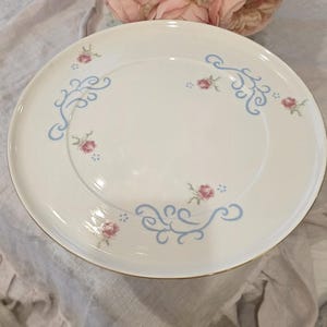 May include: White ceramic plate with a gold rim, adorned with light blue swirls and small pink rose accents. A bouquet of pink roses sits in the background, suggesting a vintage or floral theme.