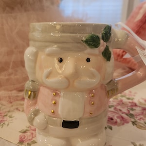 May include: A ceramic mug shaped like a nutcracker soldier. The mug features a pink jacket, white shirt, and a gray hat. The handle is pink, and the mug is decorated with gold accents and green leaves.