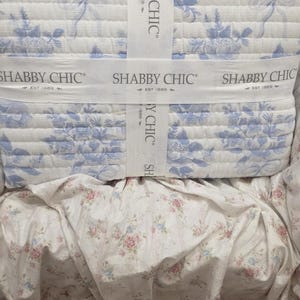 May include: A folded quilt with a blue floral pattern, secured with a white ribbon that reads "SHABBY CHIC EST 1989". The quilt is layered over a floral patterned bed skirt.
