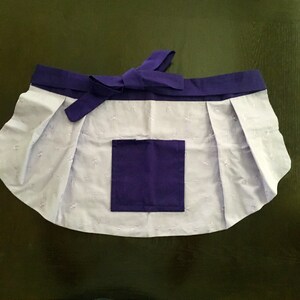 May include: A purple and white apron with a large pocket on the front. The apron has a gathered skirt and a tie at the waist.
