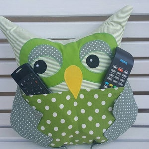 May include: A green owl-shaped pillow with a white and green polka dot pocket on the front. Two black remote controls are in the pocket.