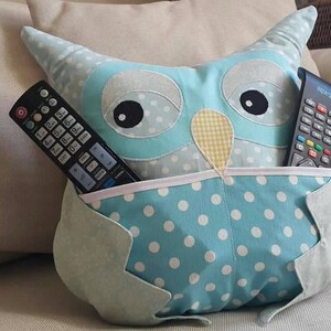 May include: A blue owl-shaped pillow with white polka dots and a pocket on the front. Two black remote controls are in the pocket.