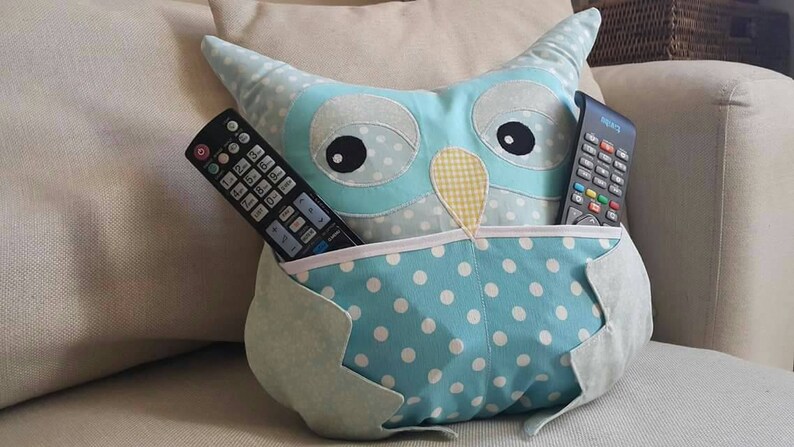 May include: A blue owl-shaped pillow with white polka dots and a pocket on the front. Two black remote controls are in the pocket.