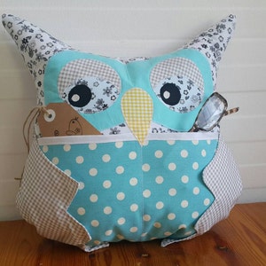 May include: A blue and white owl-shaped pillow with a polka dot pattern and a pocket for glasses. The owl has a brown tag with a white design on it.