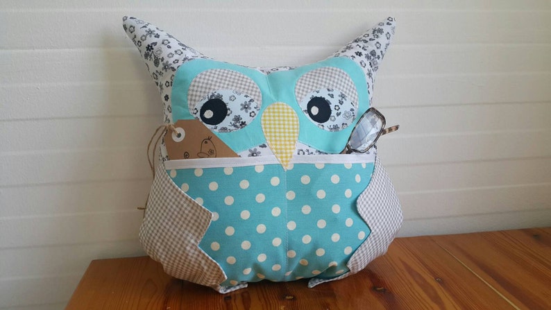 May include: A blue and white owl-shaped pillow with a polka dot pattern and a pocket for glasses. The owl has a brown tag with a white design on it.
