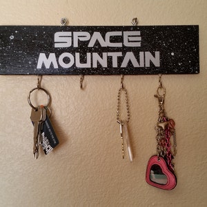 May include: A black wooden key holder with the text "SPACE MOUNTAIN" in white lettering. The key holder has five hooks and is decorated with white dots to resemble stars. There are keys hanging from three of the hooks.