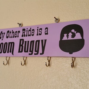 May include: A lavender wooden sign with the text "My Other Ride is a Doom Buggy" in black. A black silhouette of a Doom Buggy ride vehicle is on the right. Four silver hooks are below the sign for hanging items.