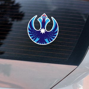 Puede incluir: Una pegatina redonda con un emblema azul, blanco y rojo, adherida a la luneta trasera de un coche. El diseño es un pájaro estilizado con una estrella en el centro. La pegatina tiene un borde blanco.