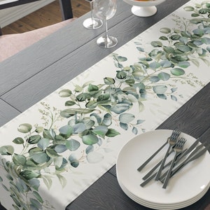 May include: A rectangular table runner with a watercolor eucalyptus leaf design in shades of green and gray. The runner is placed on a dark wooden table set for a meal, with plates, silverware, wine glasses, and a bowl of apples.