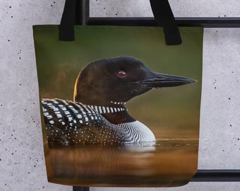 May include: A black and white loon with red eyes is swimming in a lake. The loon is in the foreground of the image, and the background is a blurry green and brown.