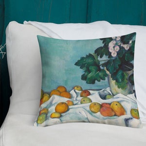 Puede incluir: Una almohada de tiro con una pintura de bodegón de un mantel blanco con una variedad de naranjas y manzanas, una planta en maceta con hojas verdes y flores blancas, y un fondo azul.