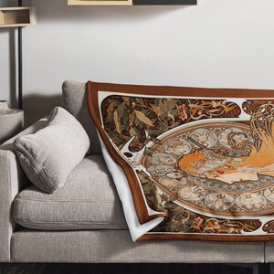 Könnte beinhalten: Eine dekorative Wohndecke mit einer Illustration im Jugendstil, die ein Frauengesicht und fließendes Haar zeigt, umgeben von Tierkreiszeichen. Die Decke hat einen braunen Rand und eine cremefarbene Rückseite, drapiert über einem grauen Sofa.
