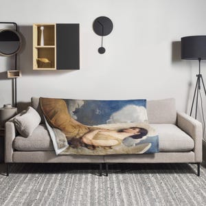 May include: A throw blanket displaying a classical painting of a woman reclining on a cloud, set against a blue sky and white clouds. The blanket is draped over a light grey sofa, bringing art into the home.