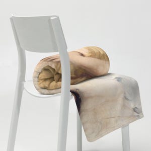May include: A rolled-up blanket in shades of beige and brown sits on a white chair. The blanket has a soft, textured appearance, with a watercolor-like design. The chair is a simple, modern design.