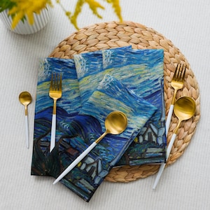 Può includere: Un set di quattro tovaglioli di stoffa blu e gialli a tema Notte stellata, forchette e cucchiai color oro con manici bianchi e un sottobicchiere intrecciato. I tovaglioli presentano un villaggio sotto un cielo notturno vorticoso.