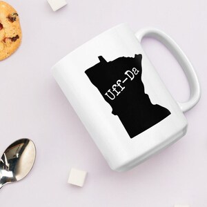 Könnte beinhalten: Weiße Keramiktasse mit einer schwarzen Silhouette des Bundesstaates Minnesota und dem aufgedruckten Text "Uff-Da".