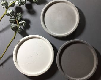 May include: Three round concrete coasters in different shades of gray. The coasters are smooth and have a slightly textured surface.