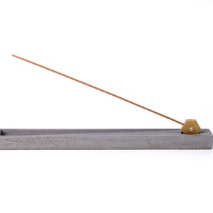 May include: A gray concrete incense holder with a brass incense stick holder. The incense stick is positioned horizontally over the holder.