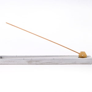 May include: A gray concrete incense holder with a brass incense stick holder. The incense stick is made of wood and is resting on the holder.