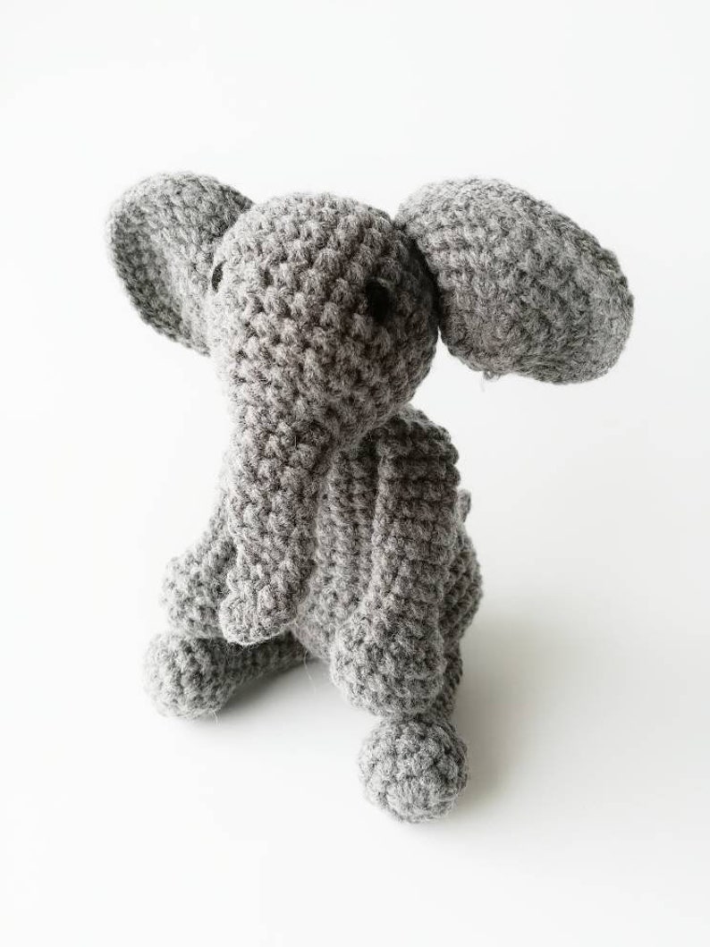 Op de afbeelding: Een grijs gehaakt olifantenspeelgoed met een kleine, opgerolde slurf. De olifant heeft zwarte ogen en zit op een wit oppervlak.
