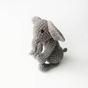 Op de afbeelding: Een grijs gehaakt knuffel olifantje met zwarte ogen. De olifant zit op een wit oppervlak.