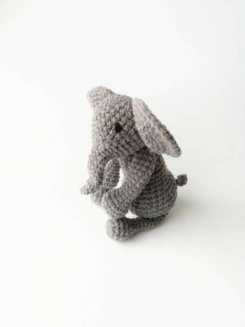 Op de afbeelding: Een grijs gehaakt knuffel olifantje met zwarte ogen. De olifant zit op een wit oppervlak.