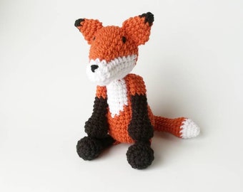 May include: A crocheted fox plush toy with orange fur, white chest, and black legs and ears. The fox is sitting with its tail curled behind it.