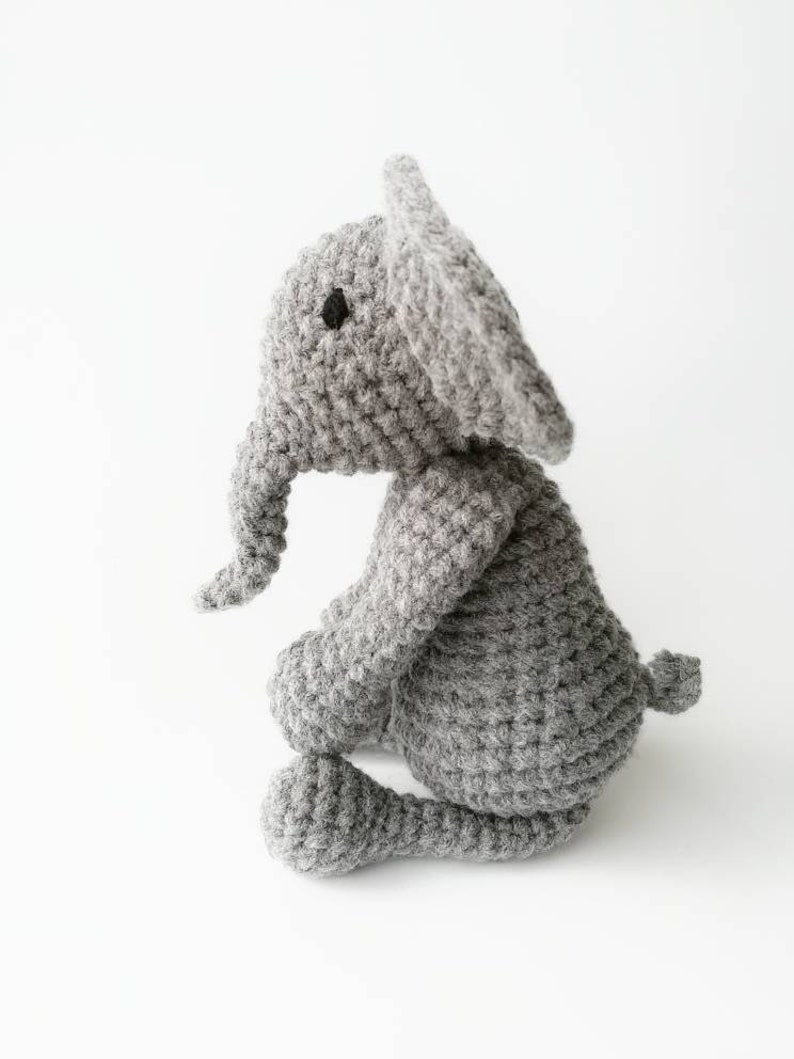 Op de afbeelding: Een grijs gehaakt knuffel olifantje dat op een wit oppervlak zit. De olifant heeft een lange slurf en grote oren.