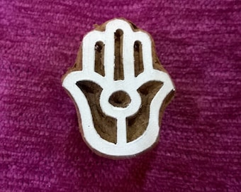 Può includere: Un blocco di legno bianco intagliato a forma di mano di hamsa, un simbolo di protezione e buona fortuna. La hamsa è un motivo popolare nelle culture del Medio Oriente e del Nord Africa.