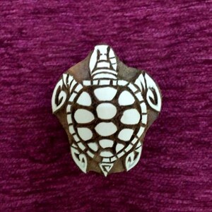 May include: A wooden stamp with a turtle design. The turtle has a white shell with oval shapes and a brown wooden frame. The stamp is on a purple background. The stamp is likely used for printing or crafting.