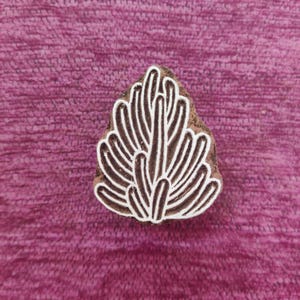 Puede incluir: Un sello de impresión de madera con un diseño blanco de una planta estilizada con hojas largas y delgadas. El sello es marrón y tiene una textura rugosa.