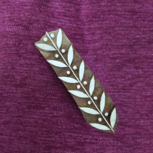Könnte beinhalten: Ein rechteckiger Holzblock mit einem Blattdesign. Der Block ist braun mit weißen Blatt- und Punktmustern. Der Hintergrund ist ein strukturierter, tiefvioletter Stoff.