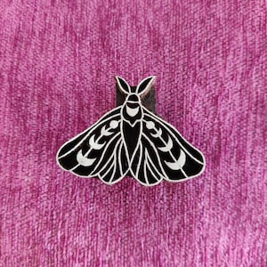 May include: A black and white moth-shaped wooden stamp. The stamp features a detailed design with white accents on the wings and body. The stamp is set against a textured pink background.