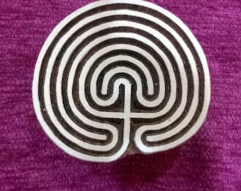 May include: A white wooden stamp with a black circular design of a labyrinth. The stamp is on a purple background.