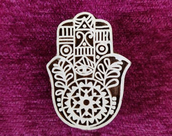 Può includere: Un blocco di legno intagliato a mano con un design dettagliato di una hamsa, un simbolo protettivo in molte culture. La hamsa è decorata con motivi intricati e un occhio centrale, che simboleggia la protezione e la buona fortuna.