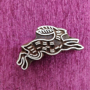 May include: A wooden block printing stamp with a detailed design of a rabbit in a running pose. The stamp is carved with intricate lines and patterns, creating a unique and decorative design.