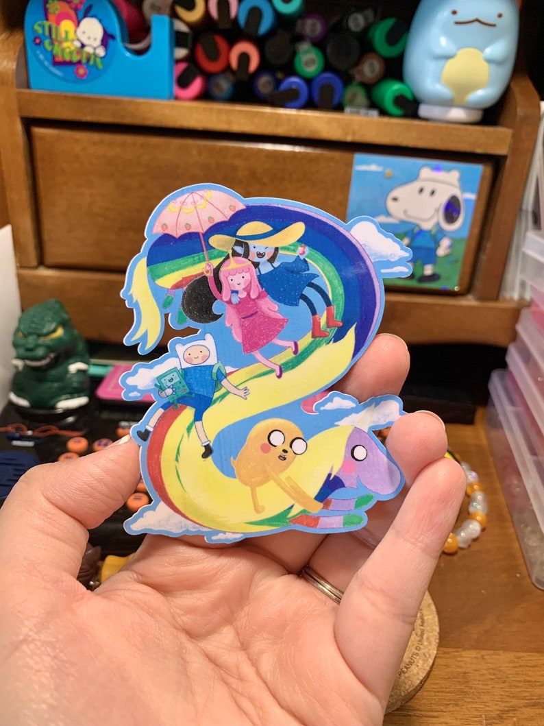 May include: A sticker featuring characters from the animated series Adventure Time. The sticker shows Finn, Jake, Princess Bubblegum, and Marceline the Vampire Queen riding a rainbow. The sticker is on a hand.