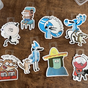Op de afbeelding: Een verzameling van acht gestanste vinylstickers met personages uit de animatieserie Regular Show. De stickers bevatten Pops met de tekst jolly good show, Rigby, Mordecai, Muscle Man met een sombrero en Benson met de tekst you're fired. Elke sticker heeft een doorzichtig plastic lipje aan de bovenkant om op te hangen.