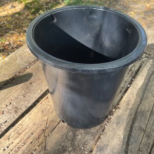 May include: A black plastic plant pot with a wide rim. The pot is cylindrical with a slightly tapered shape and has a drainage hole at the bottom. The pot is sitting on a weathered wooden surface.