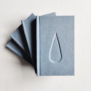 Op de afbeelding: Een stapel blauwgrijze notitieboekjes met een verdiept traanvormig ontwerp op de kaft. De notitieboekjes zijn uitgespreid, waardoor de randen van de pagina's zichtbaar zijn. Het papier heeft een textuur.