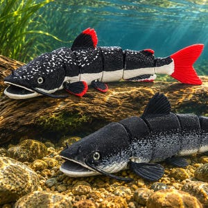 Può includere: Due statuette di pesce articolate con corpi segmentati. Il pesce in alto è nero con macchie bianche, pinne rosse e coda rossa. Il pesce in basso è nero con macchie bianche e ha pinne e coda nere. Entrambi hanno la bocca aperta.