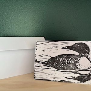 Peut inclure: Une carte de vœux blanche présentant une illustration en impression à l'encre noire d'un plongeon nageant sur l'eau, accompagnée d'une enveloppe blanche assortie. L'œuvre présente des textures contrastées et des lignes audacieuses.
