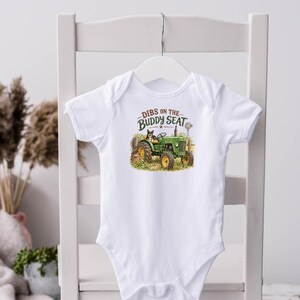 May include: A white short-sleeve baby bodysuit featuring a graphic print of a green tractor with a dog sitting on the seat. The text above the tractor reads DIBS ON THE BUDDY SEAT in dark green lettering. The garment has a round neckline and snaps.