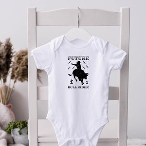 May include: White baby onesie with a black silhouette of a cowboy riding a bull. The text "FUTURE BULL RIDER" is printed below the image.