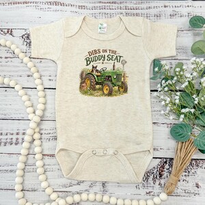 May include: A heathered beige short-sleeve baby bodysuit featuring a graphic of a green tractor with a dog sitting in the driver seat. The text above the tractor reads DIBS ON THE BUDDY SEAT in dark green and brown lettering with a small windmill.